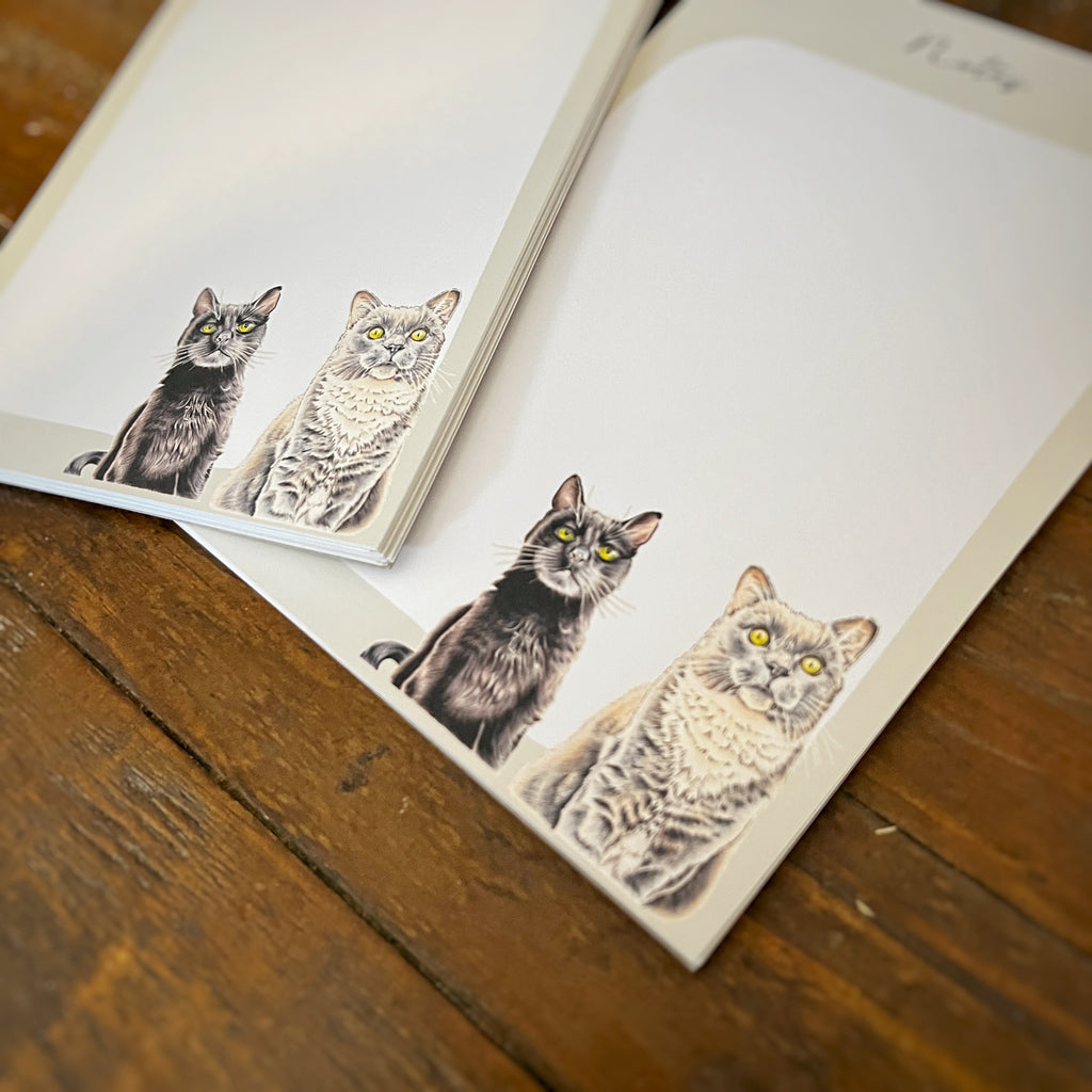 A stack of white stationery notepads featuring printed illustrations of cats, arranged on a rustic wooden tabletop.