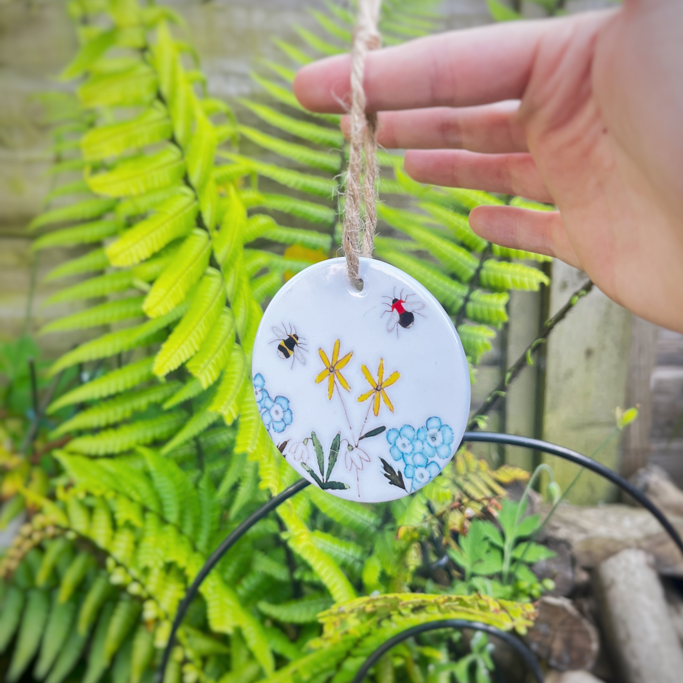 A round white ceramic hanging ornament decorated with colorful painted flowers, bees, and a ladybug, suspended by a twine string. A human hand is holding the ornament in front of a background of green ferns and garden foliage.