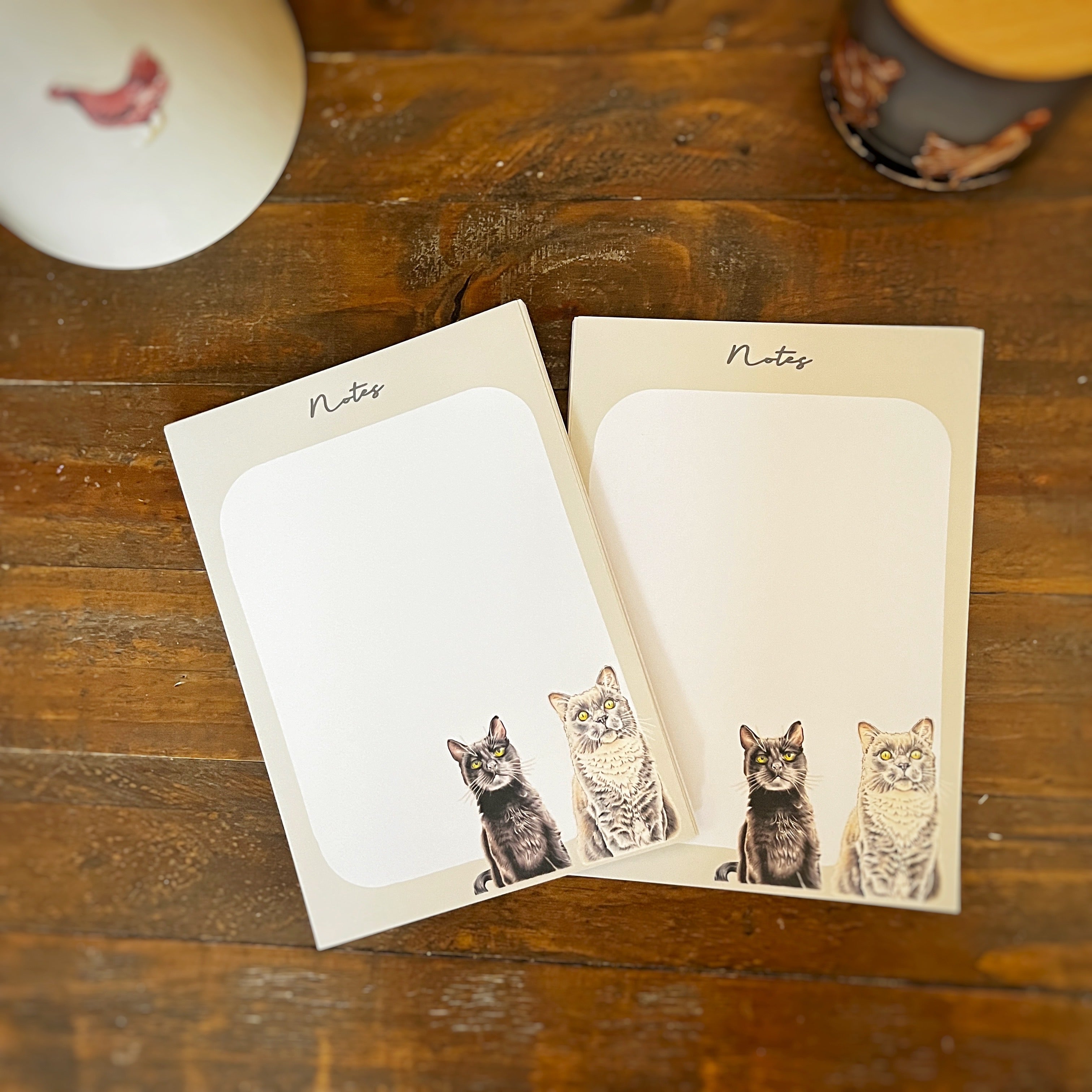 Two cream-colored notepad sheets with rounded white writing areas and printed cat illustrations at the bottom, placed on a wooden tabletop. The word 'Notes' appears at the top of each pad.