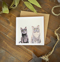 A square greeting card featuring a hand drawn illustration of two cats, one black cat and one grey cat placed on top of a kraft brown envelope over a rustic wooden surface with dried leaves and twine.