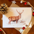 A rectangular art greeting card featuring a brown stag with antlers standing in a snowy landscape, styled on a wooden surface with kraft envelopes, rope, pinecones, and red jingle bells.