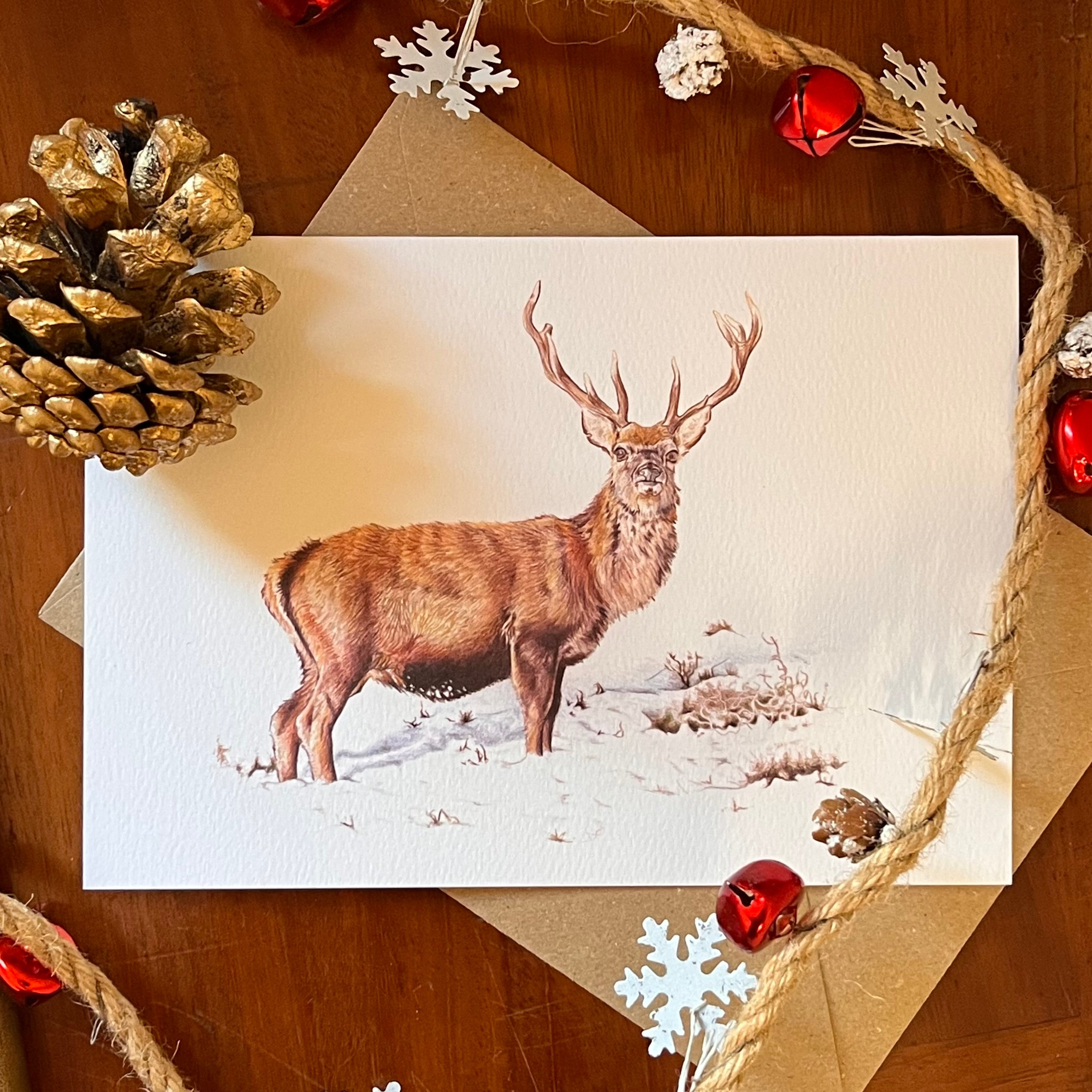 A rectangular art greeting card featuring a brown stag with antlers standing in a snowy landscape, styled on a wooden surface with kraft envelopes, rope, pinecones, and red jingle bells.