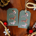 Two rectangular Christmas gift tags with rounded corners, printed with illustrated dachshunds wearing Santa hats, laid on a wooden surface with twine and holiday decorations around them.