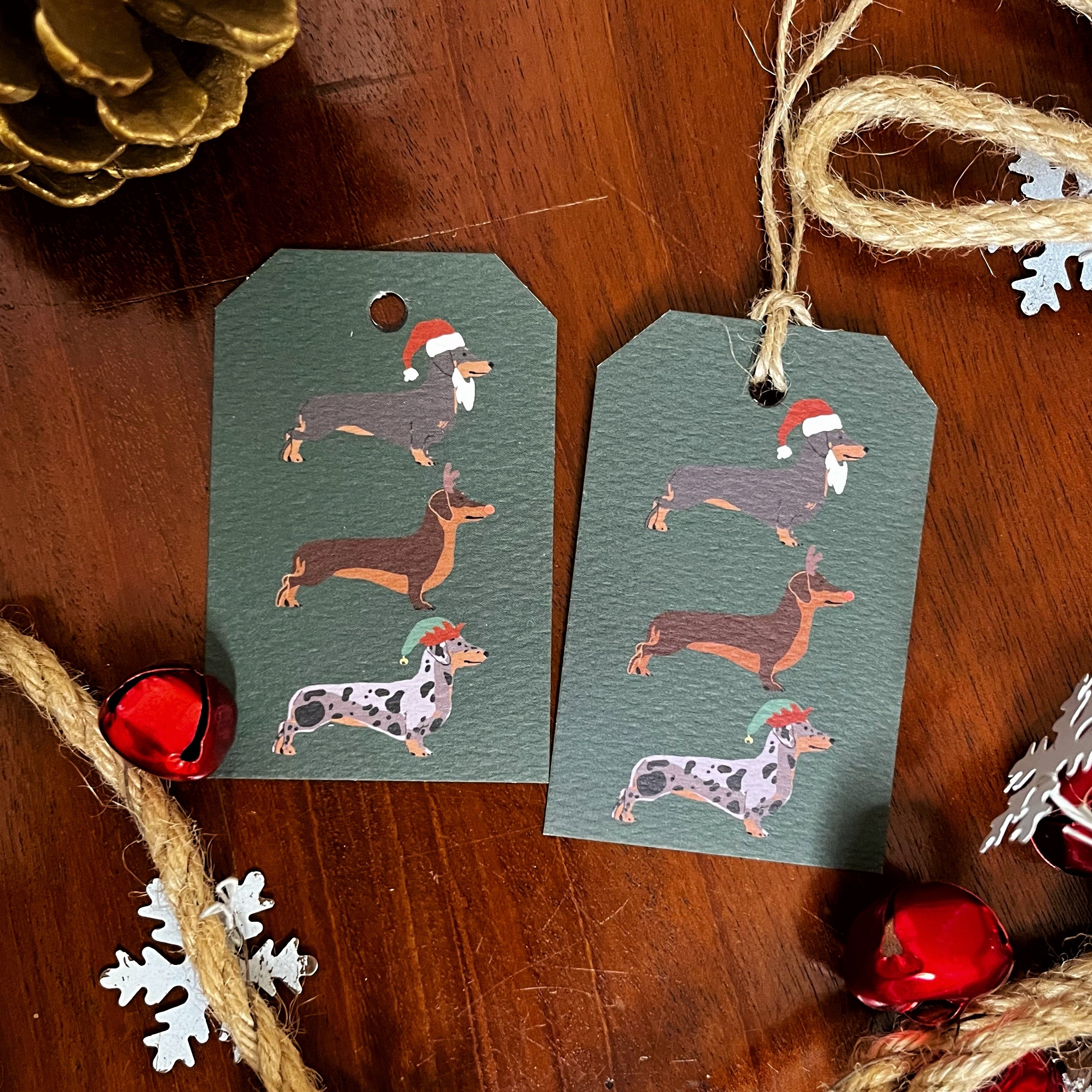 Two rectangular Christmas gift tags with rounded corners, printed with illustrated dachshunds wearing Santa hats, laid on a wooden surface with twine and holiday decorations around them.