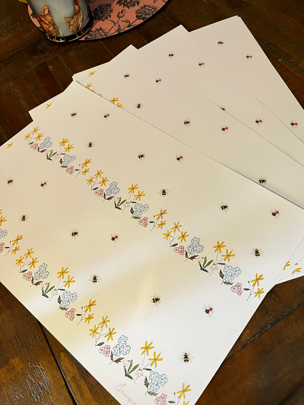 A stack of white wrapping paper sheets with a delicate floral and bee pattern, displayed on a dark wooden table.