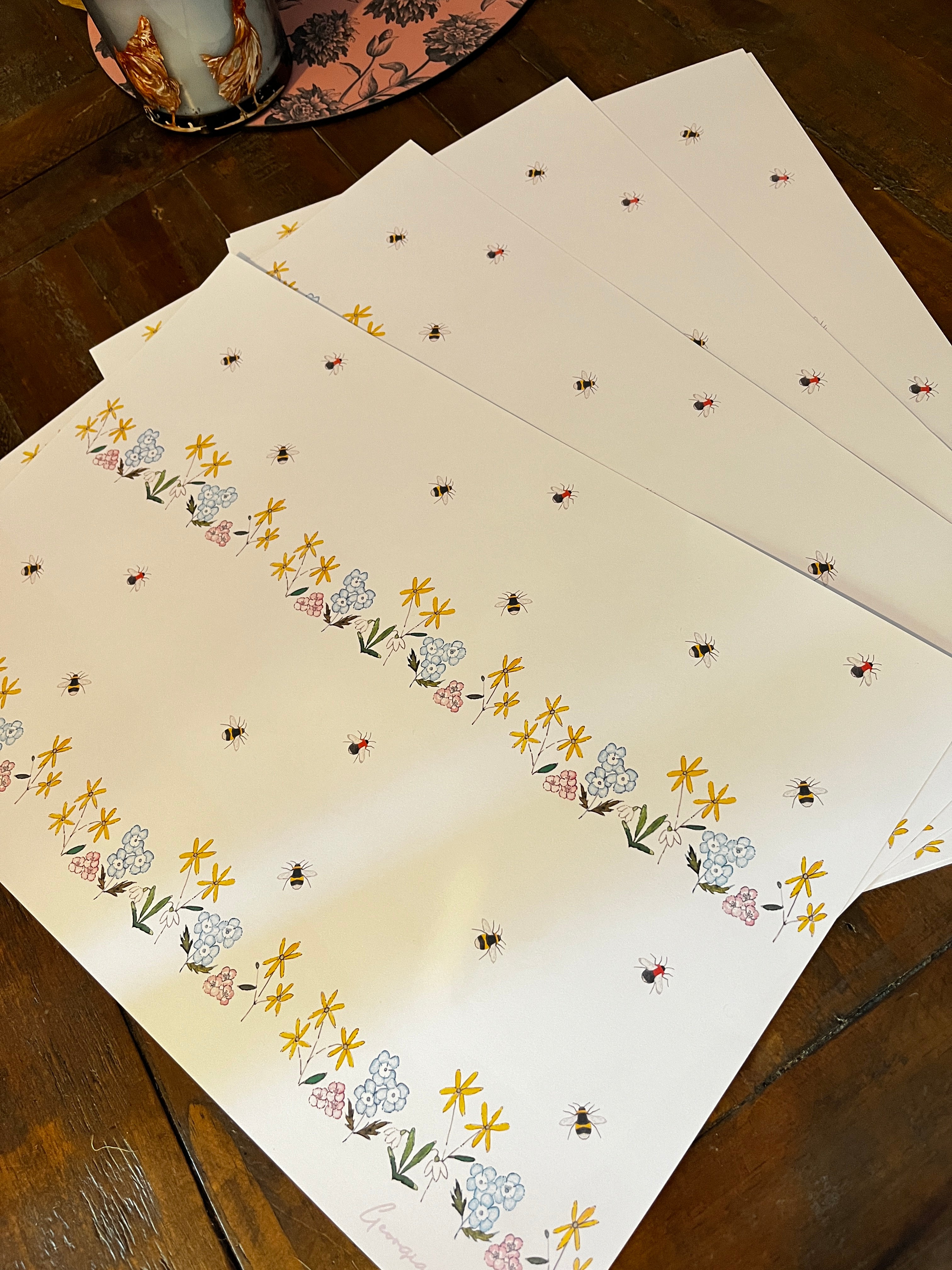A stack of white wrapping paper sheets with a delicate floral and bee pattern, displayed on a dark wooden table.