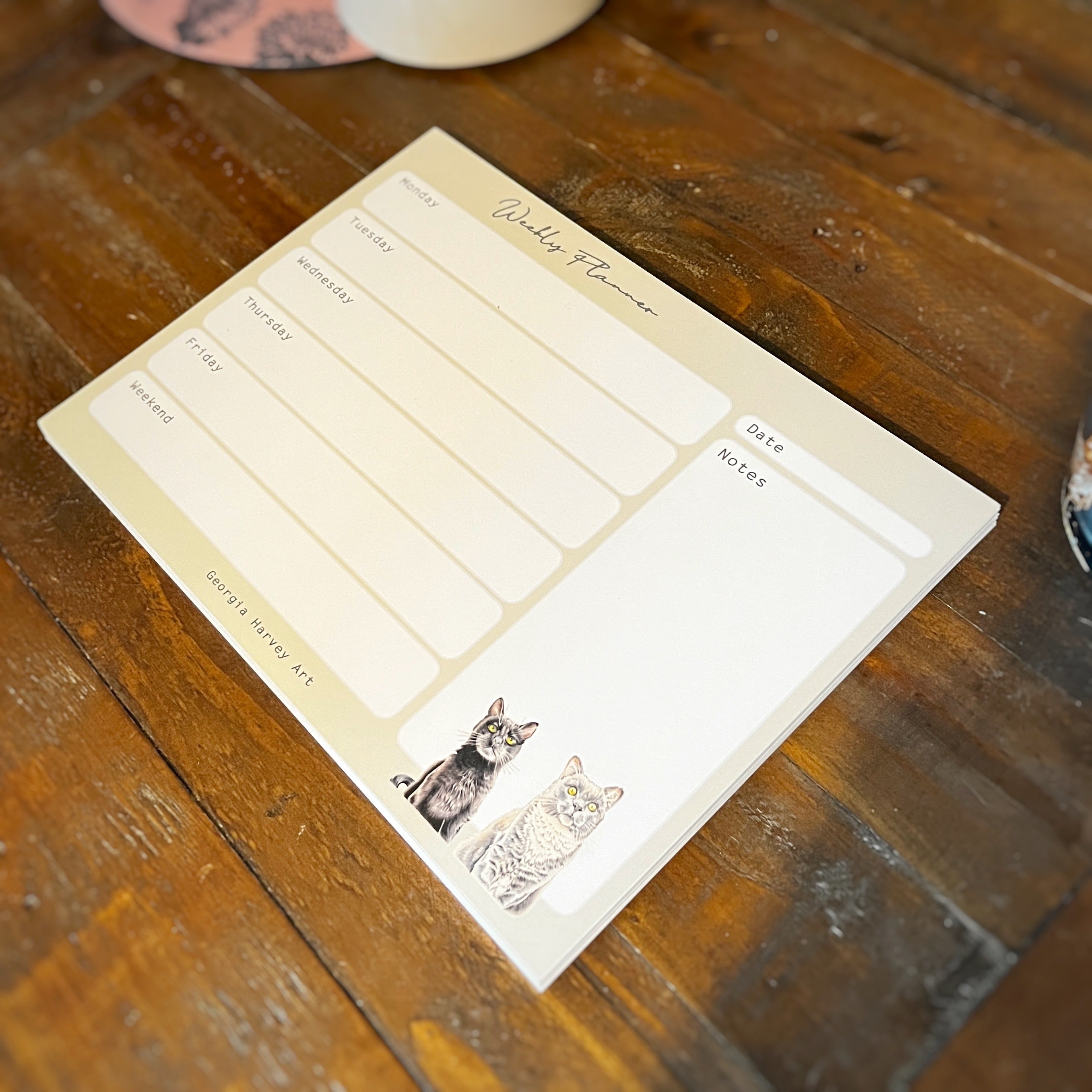 A weekly planner notepad with cute cat illustrations, resting on a rustic wooden table.