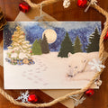 A watercolor-style Christmas card featuring a snowy winter night scene with a polar bear in the snow, evergreen trees, a decorated tree, gift boxes, and a full moon. The card is styled on a brown wooden surface with kraft paper, rope, and red holiday ornaments around it.