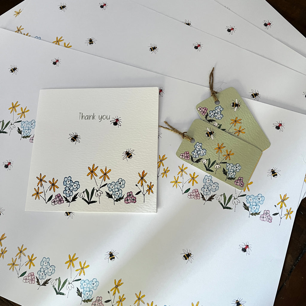 A stationery set of bumbebee with flowers illustrations. White thank you card, matching decorative tags tied with twine, arranged on a matching patterned A3 wrapping paper sheet.