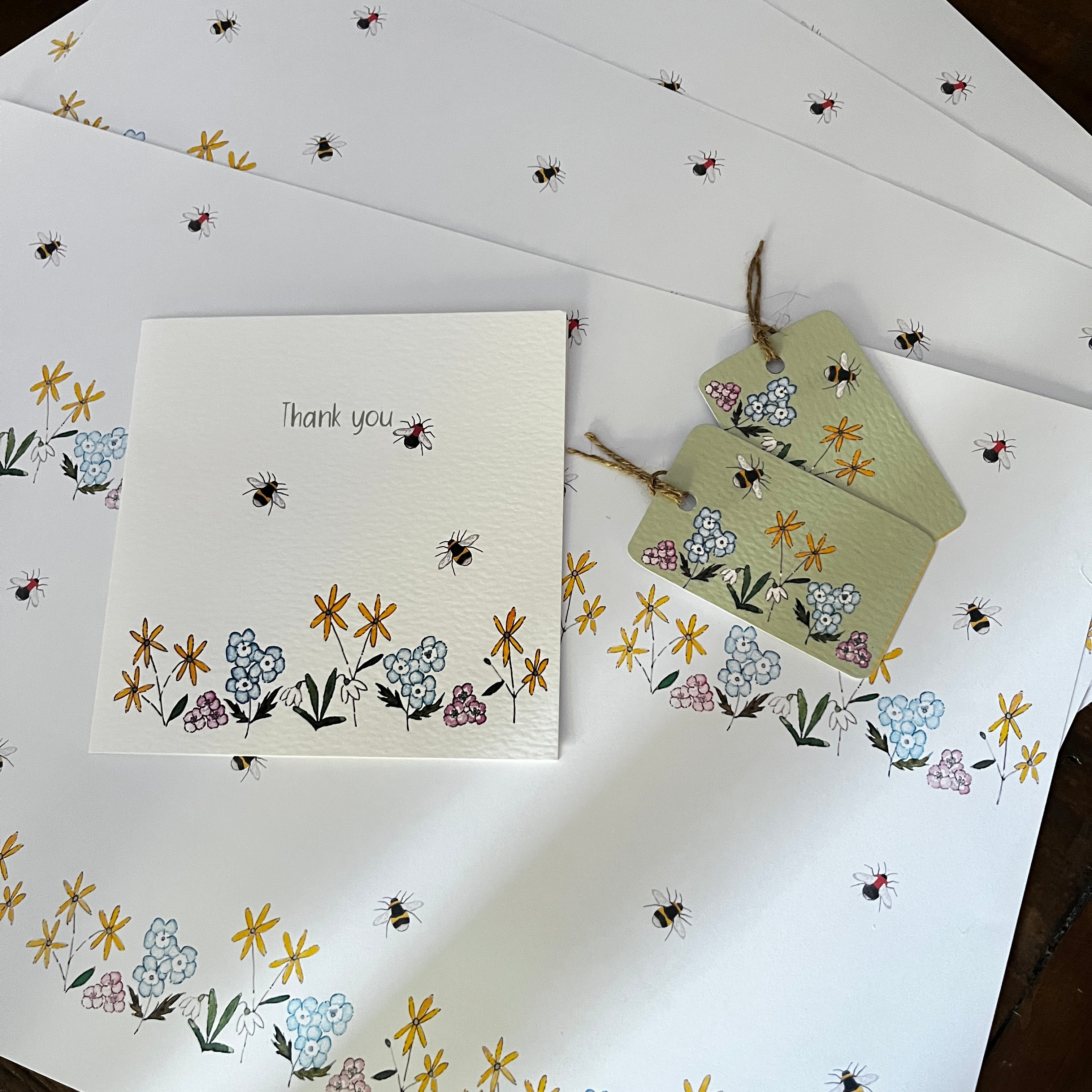 A stationery set of bumbebee with flowers illustrations. White thank you card, matching decorative tags tied with twine, arranged on a matching patterned A3 wrapping paper sheet.