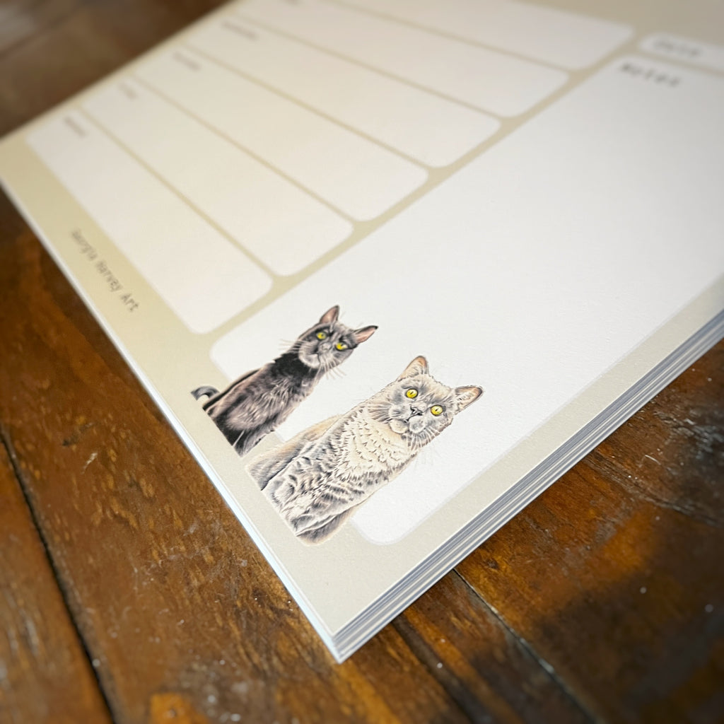 A close-up of a cream-colored notepad with two illustrated cats at the bottom, placed on a dark wooden tabletop.
