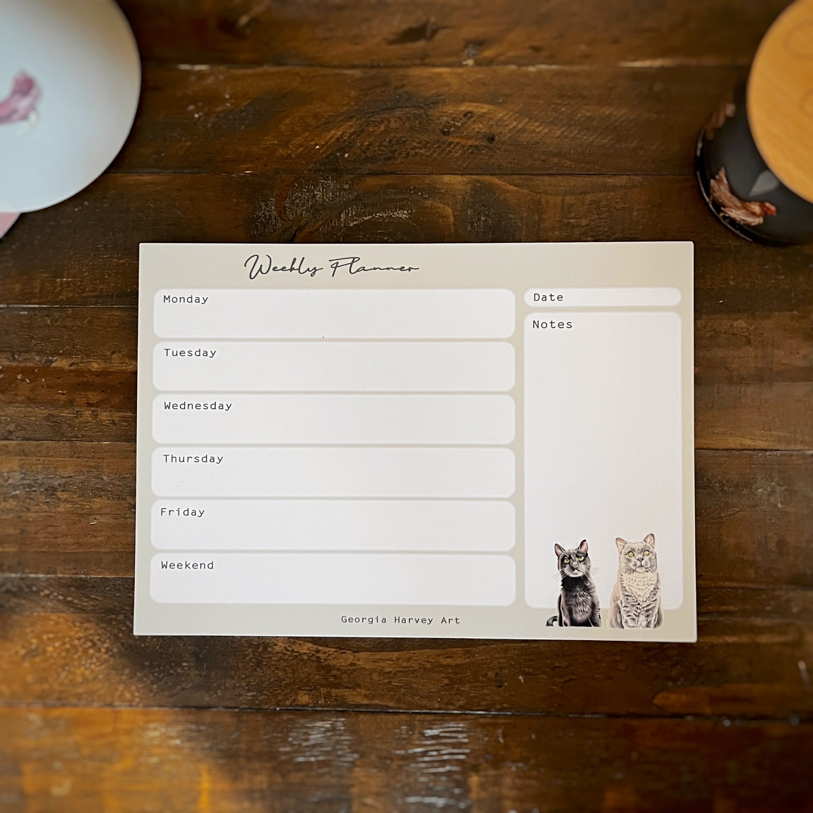 A weekly planner notepad with cute cat illustrations, laid flat on a rustic dark wood tabletop.