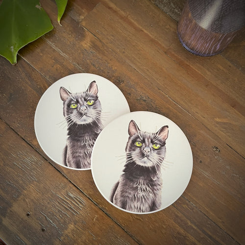 Two round coasters featuring a printed illustration of a black cat with green eyes, displayed on a rustic wooden tabletop.