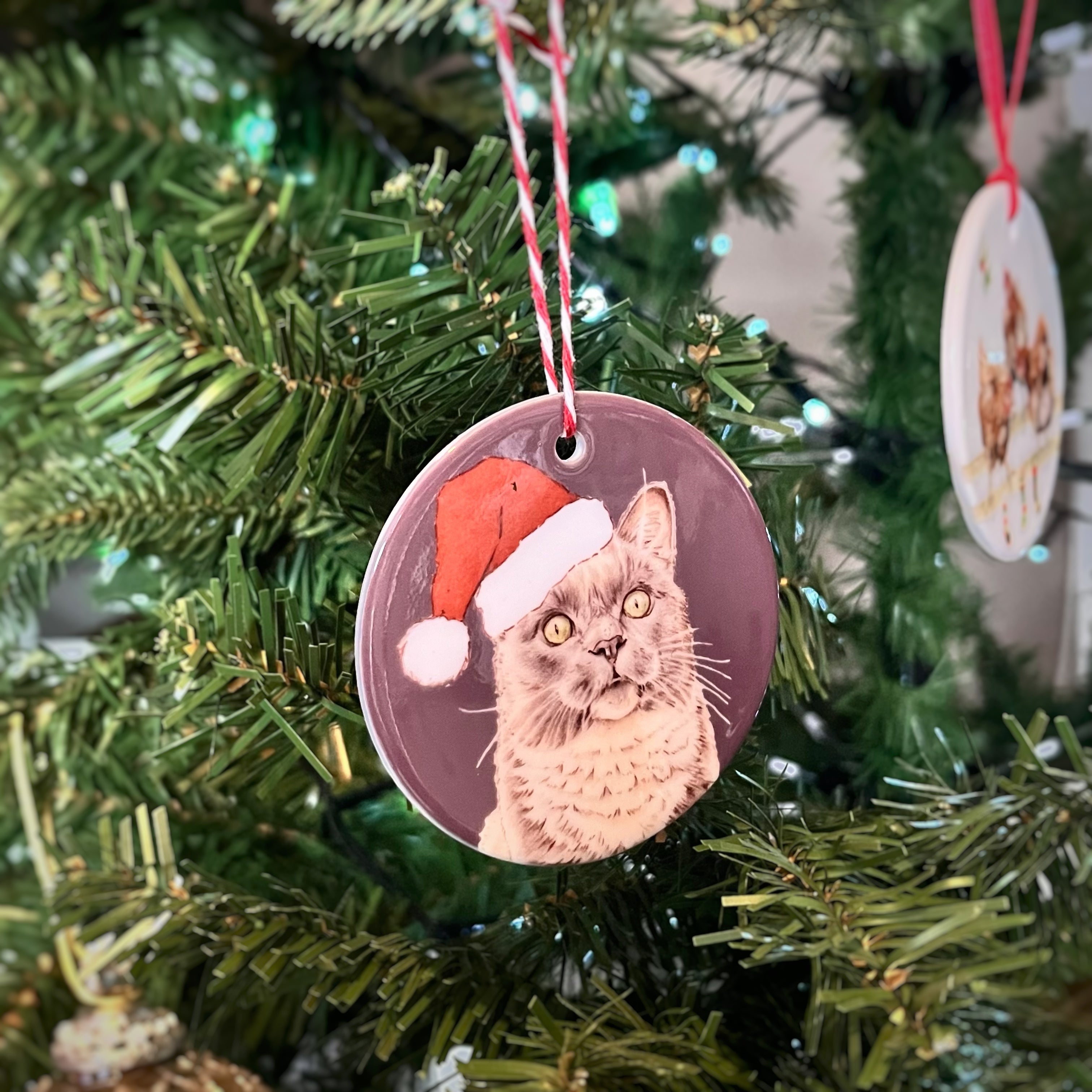 A round ceramic Christmas ornament featuring a printed illustration of a grey cat wearing a red Santa hat, hanging from a red and white string on a green Christmas tree.