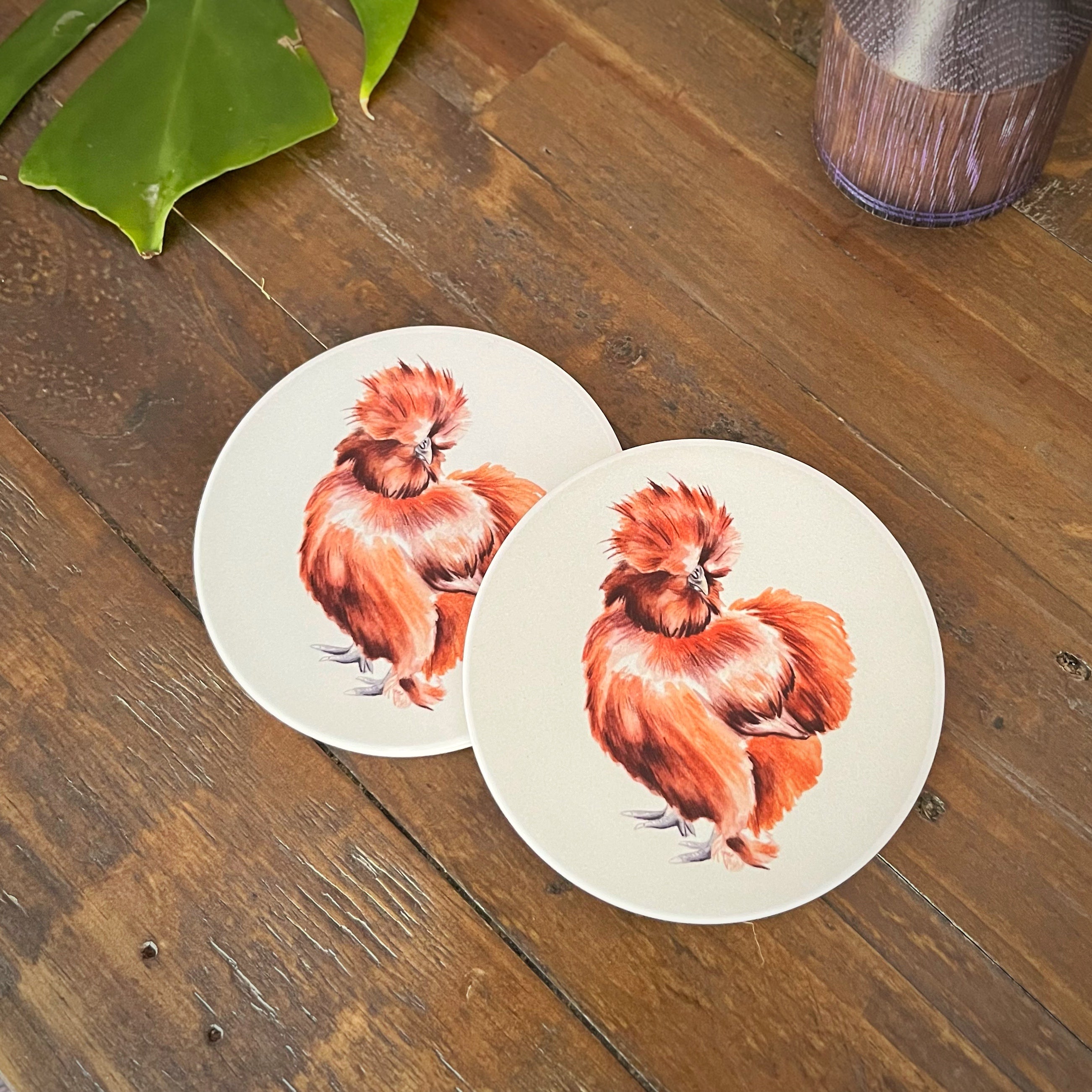 Two round ceramic coasters featuring a detailed illustration of a fluffy red silkie chicken, displayed on a rustic wooden tabletop.