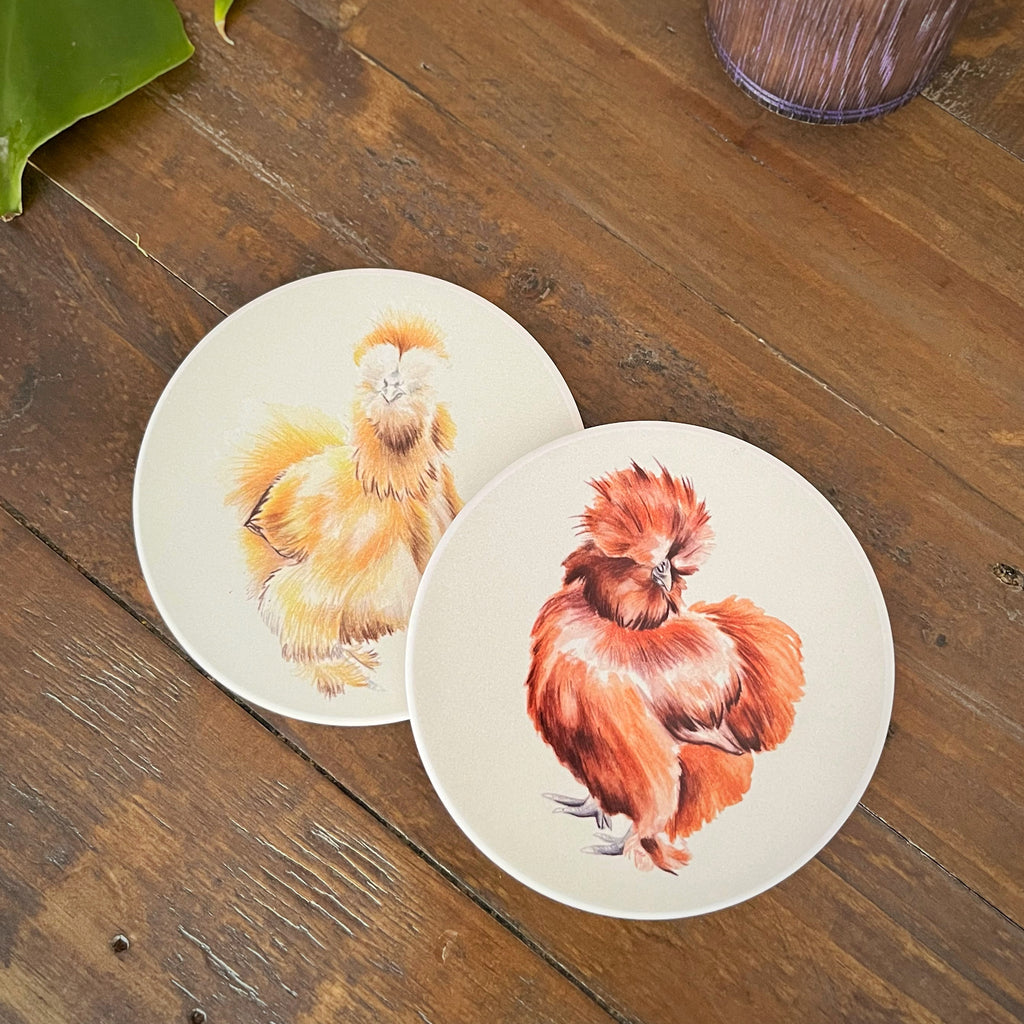 Two round coasters featuring painted chickens, displayed on a rustic wooden tabletop. One coaster shows a fluffy yellow chicken, and the other shows a reddish-brown chicken.