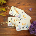 Four light green coloured gift tags with rustic twine loops, decorated with illustrations of blue, pink, and yellow flowers, bumblebees, and dandelions, arranged on a wooden surface with dried flowers.