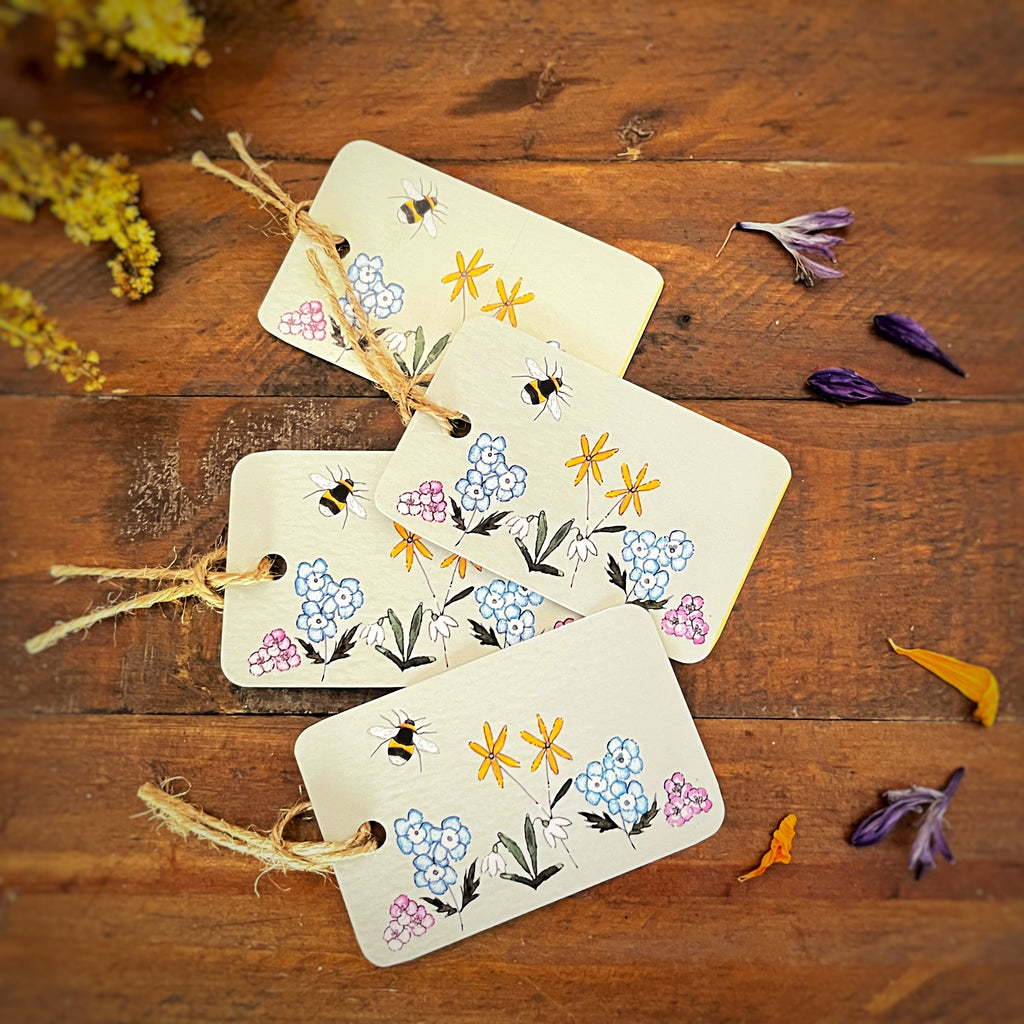 Four light green coloured rectangular gift tags with rustic twine loops, decorated with hand-painted bees, blue and yellow flowers, and green leaves, arranged on a wooden surface with dried flowers.