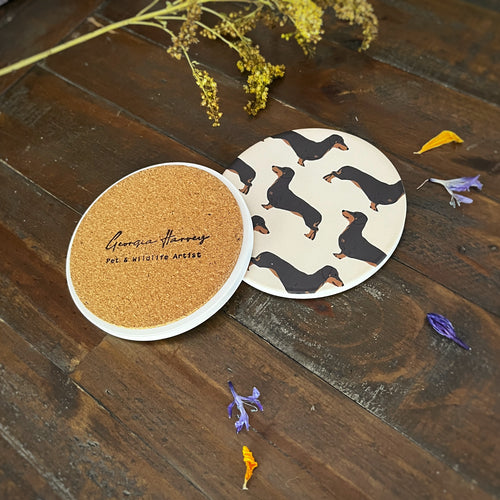 Two round coasters showing an illustration of Black and Tan sausage dogs on a dark wooden table with dried flowers. One coaster is upside down, showing a natural cork insert with the logo 'Georgia Harvey Pet & Wildlife Art' stamped in black script.