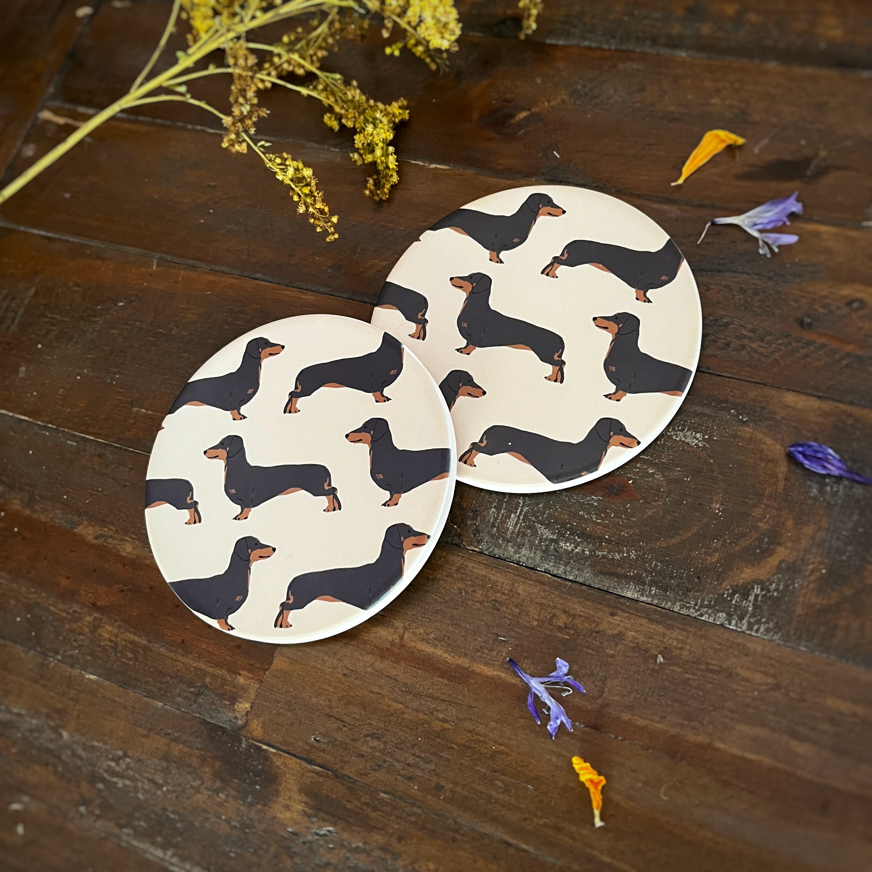 Two round coasters with a Black and Tan dachshund pattern, displayed on a rustic dark wood surface with dried flowers.