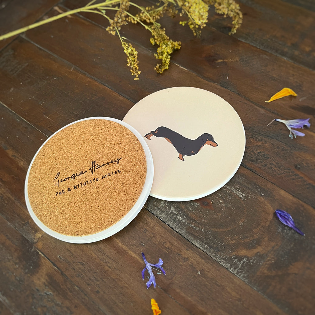Two round coasters with a Black and Tan sausage dog design on a dark wooden tabletop. One coaster has a cork backing with a stamped logo reading 'Georgia Harvey Pet & Wildlife Art'.
