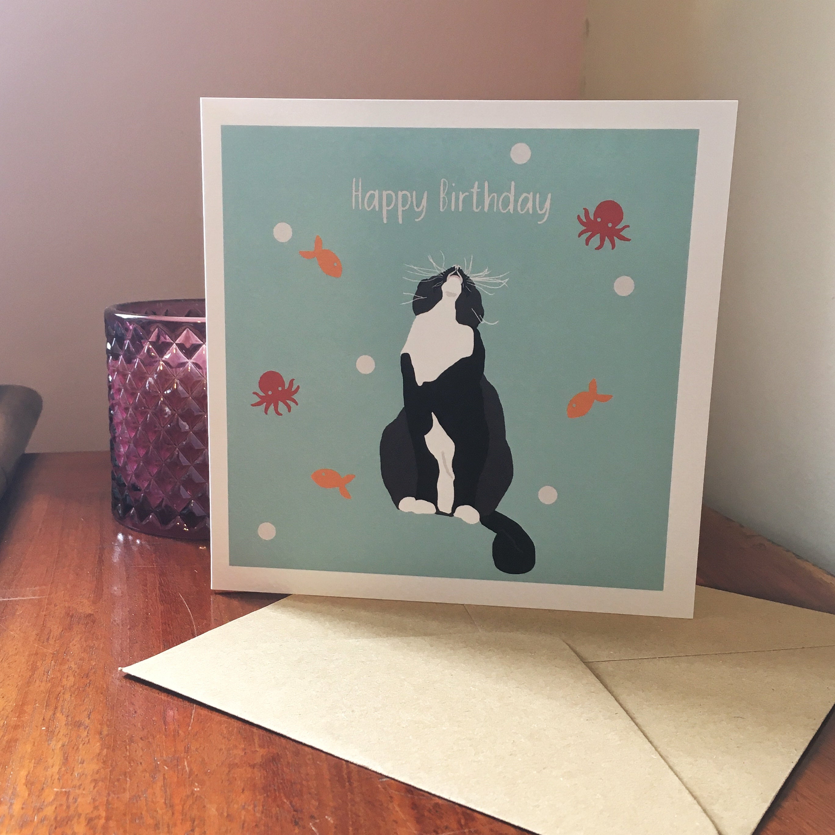 birthday card of a black and white cat looking up with a blue background, white dots, orange fish and red octopus around the cat.
