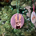 A round ceramic Christmas ornament featuring a black cat illustration with antlers, hanging on a green Christmas tree with lights. Another ornament is visible in the background.