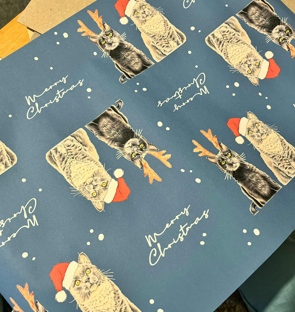 Blue wrapping paper featuring a repeating pattern of cats wearing Santa hats and reindeer antlers, with white script reading 'Merry Christmas' and snow around them.
