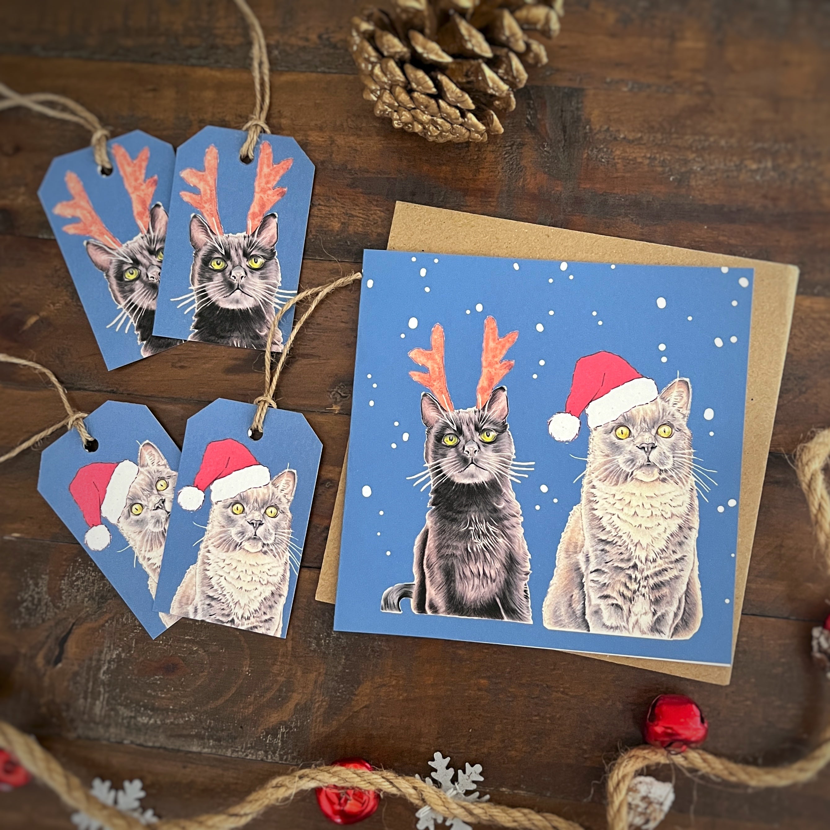 A Christmas-themed greeting card set featuring a blue card and matching tags with illustrated black cat and grey cat wearing Santa hats and reindeer antlers, arranged on a dark wooden surface with a pinecone and jingle bells as props.