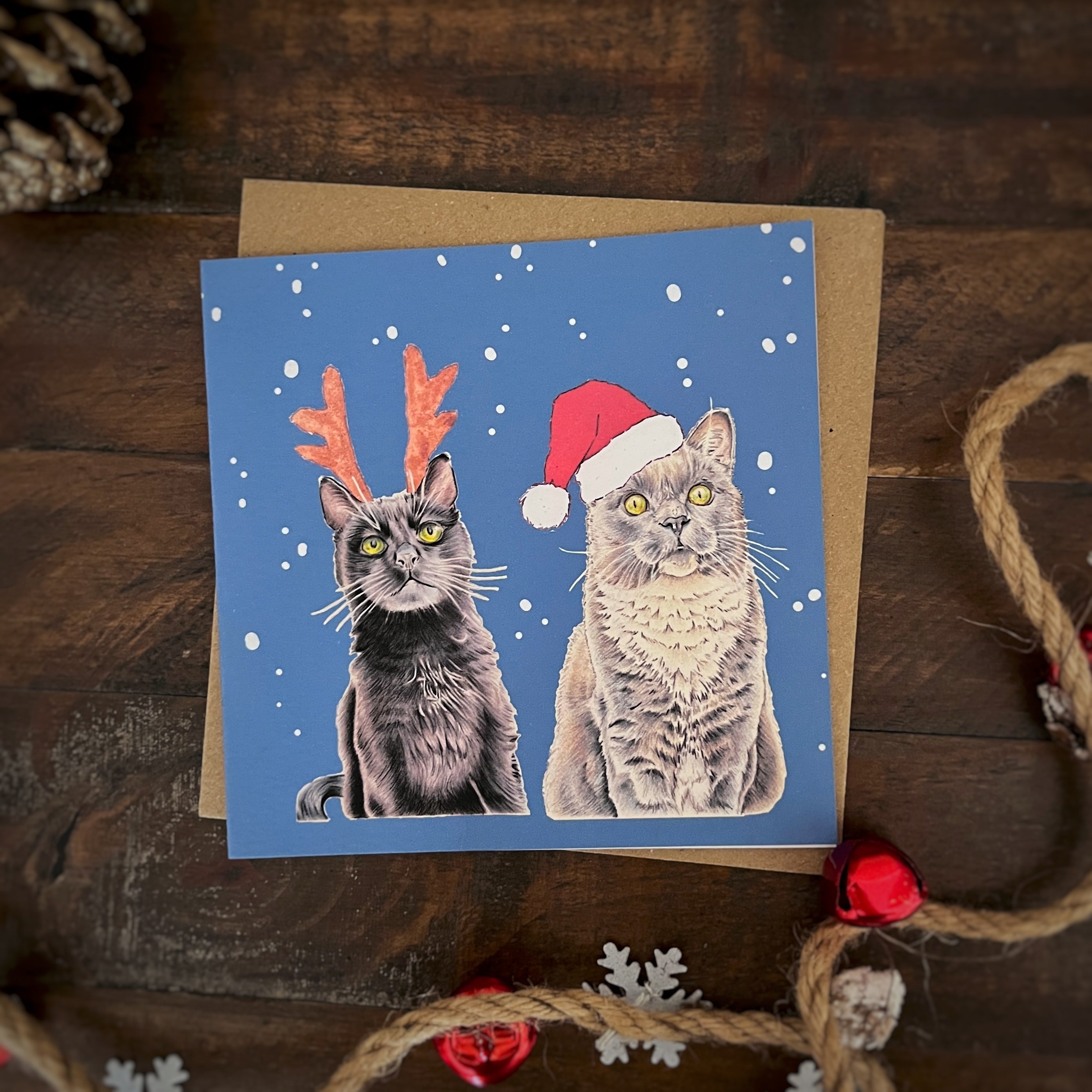 Blue Christmas-themed christmas card featuring illustrated black cat and grey cat wearing Santa hats and reindeer antlers, scattered on a wooden surface with holiday decorations including twine, a pinecone, and red ornaments.