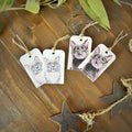 Four white rectangular gift tags printed with a gray cat and black cat portraits, hanging from natural jute twine on a rustic wooden surface with decorative leaves and star-shaped ornaments.