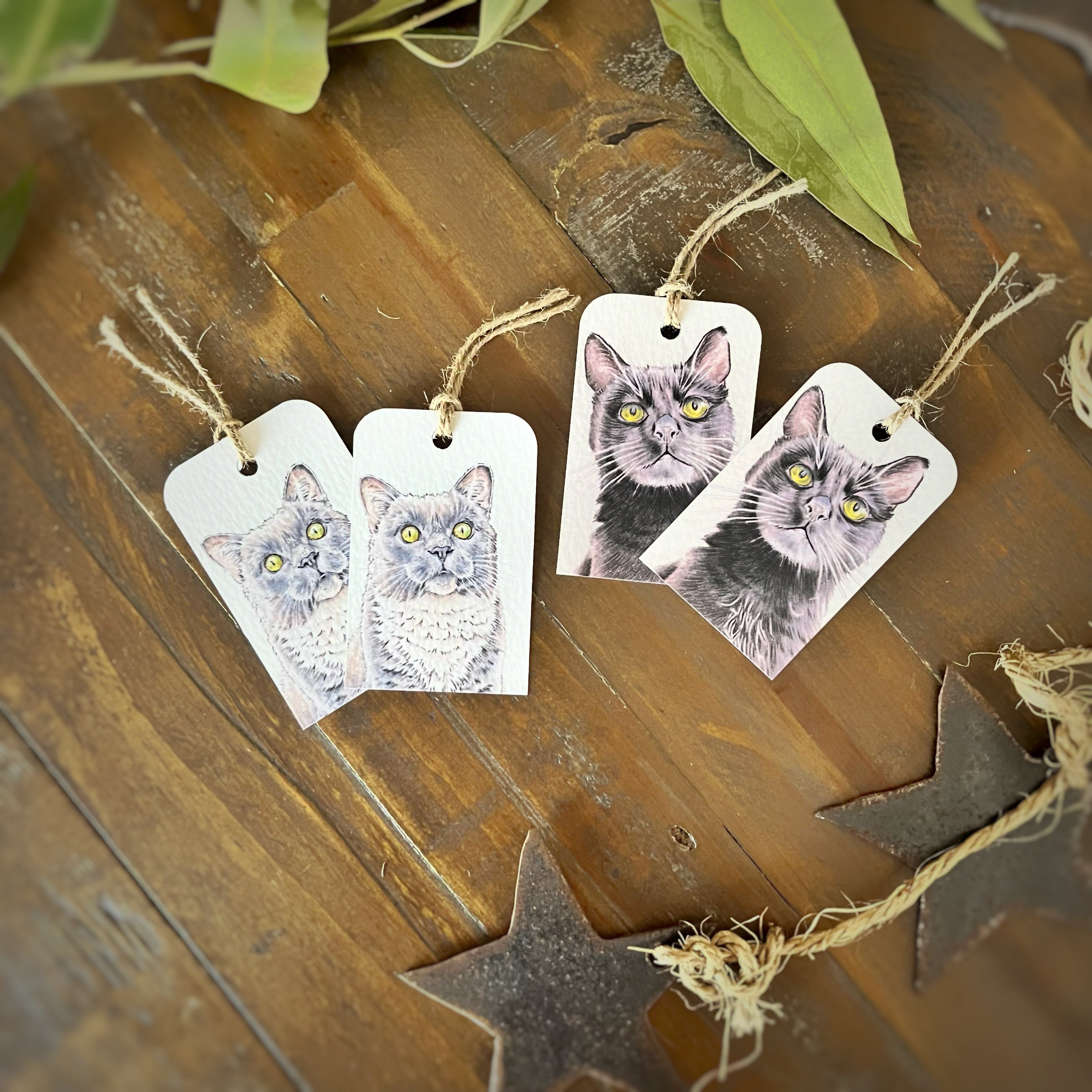 Four white rectangular gift tags printed with a gray cat and black cat portraits, hanging from natural jute twine on a rustic wooden surface with decorative leaves and star-shaped ornaments.