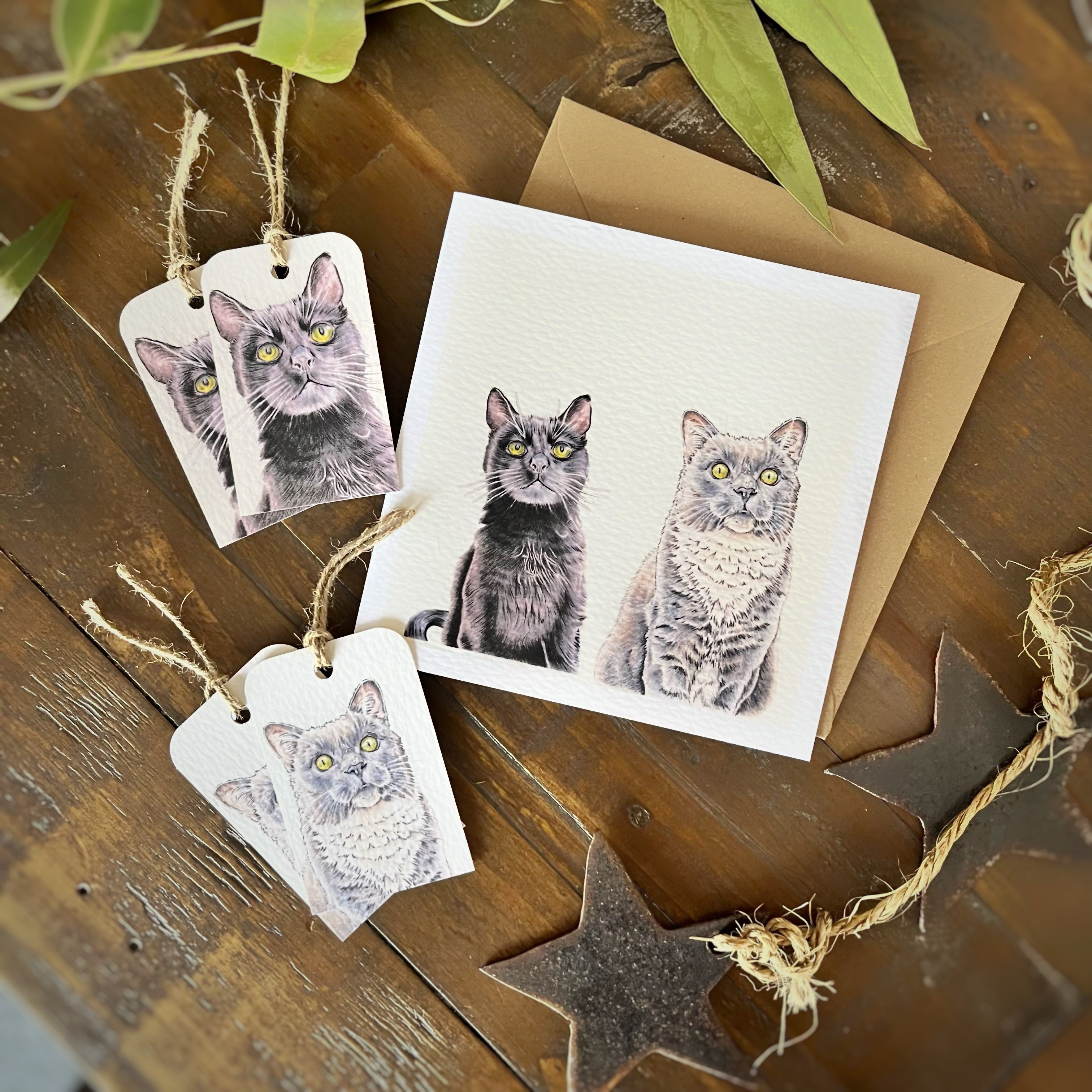 A square greeting card and kraft envelope on a rustic wooden surface, each printed with a black cat and grey cat portrait. Four small matching gift tags are also arranged around them with twine loops.