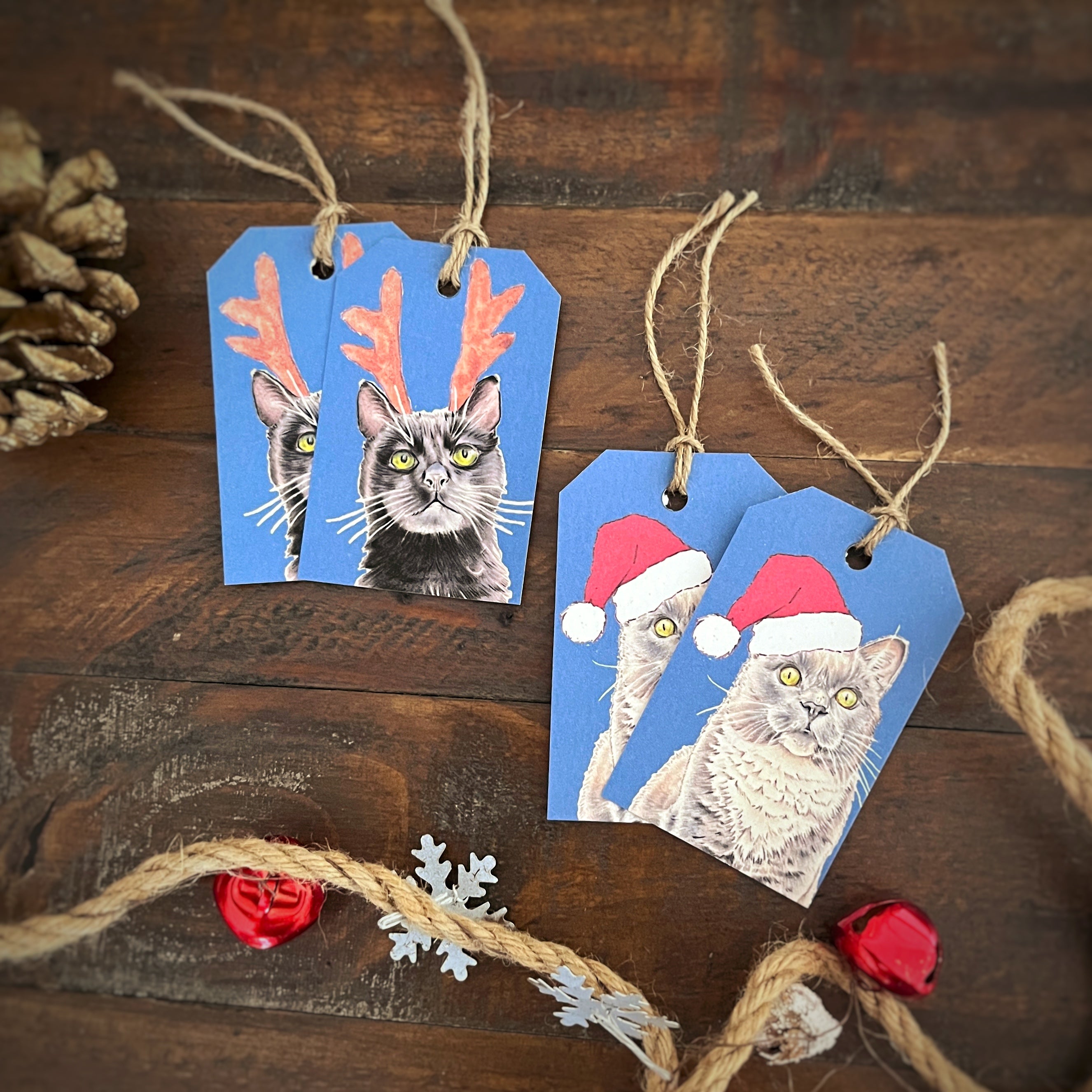 Two blue Christmas gift tags with twine, printed with cat portraits wearing festive accessories, arranged on a dark wooden surface with holiday props.