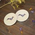 Two round coasters with a cream background, printed with a dapple dachshund illustration, displayed on a dark wooden tabletop with dried flowers and petals.