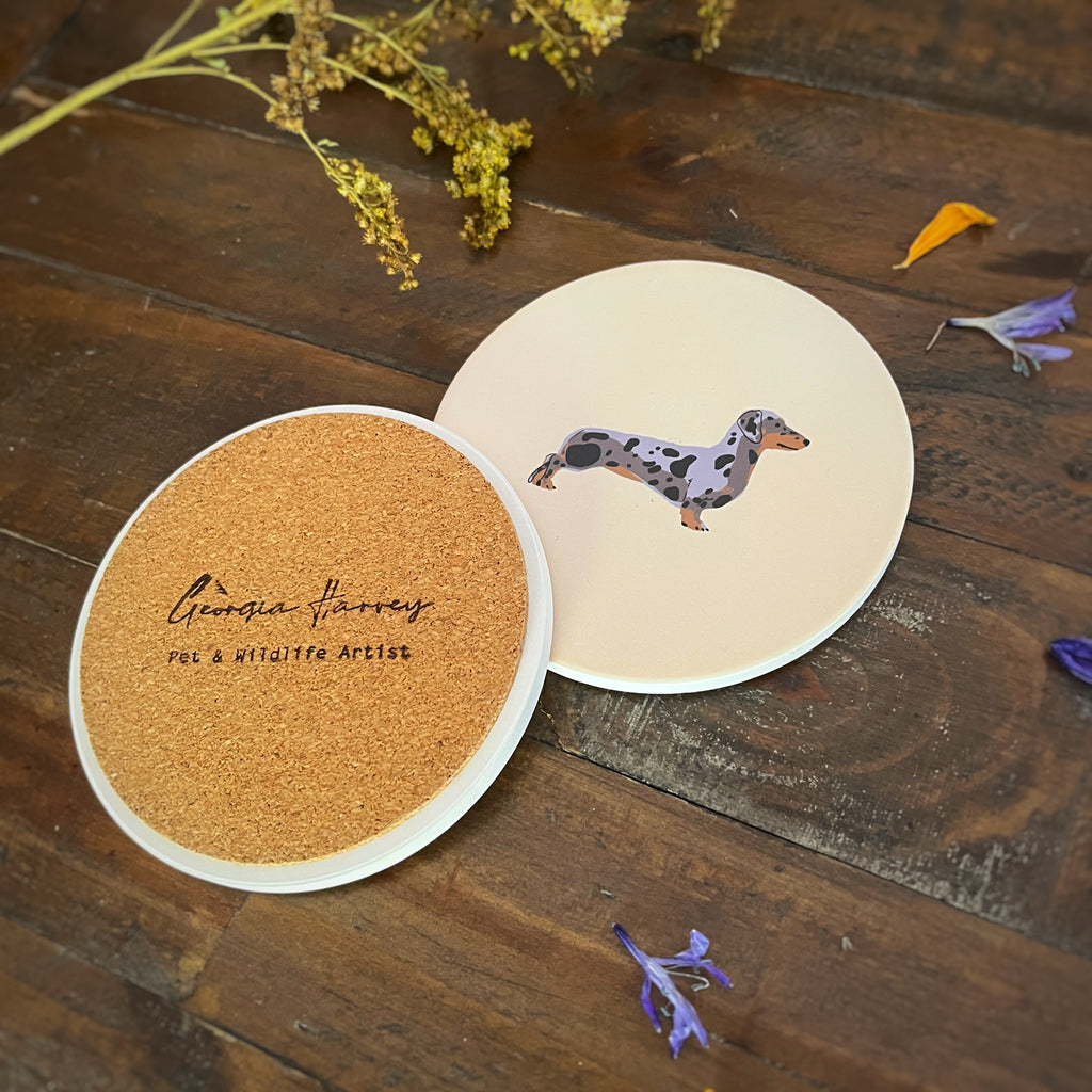 Two round coasters with an illustration of a dapple sausage dog on a rustic dark wood surface. One coaster shows a natural cork backing with the text 'Georgia Harvey Pet & Wildlife Artist' stamped on it.