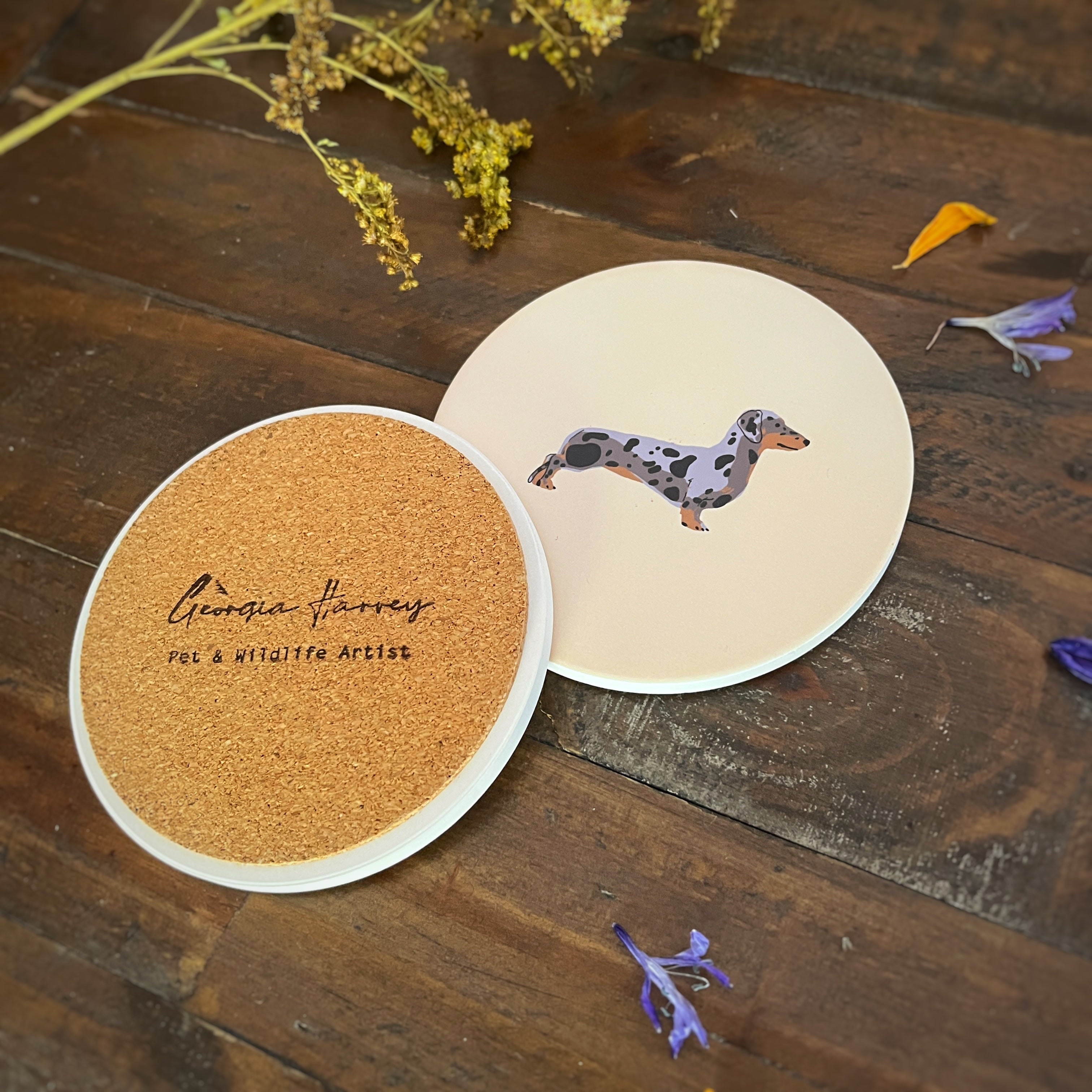 Two round coasters with an illustration of a dapple sausage dog on a rustic dark wood surface. One coaster shows a natural cork backing with the text 'Georgia Harvey Pet & Wildlife Artist' stamped on it.