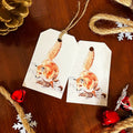 Two white paper gift tags with a rustic twine loop, printed with a watercolor illustration of a red fox in winter, displayed on a dark wooden surface with holiday decorations.