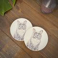 Two round coasters featuring a detailed illustration of a gray cat with green, yellow eyes, displayed on a rustic wooden tabletop.