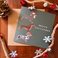 A Christmas greeting card featuring three dachshund illustrations wearing holiday hats, placed on a kraft envelope over a wooden surface with rope, snowflakes, red bells, and a pinecone around it. The card reads 'Merry Christmas'.