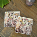 Two square coasters with a detailed illustration of a rabbit in a field of grass and wildflowers, displayed on a rustic wooden tabletop.