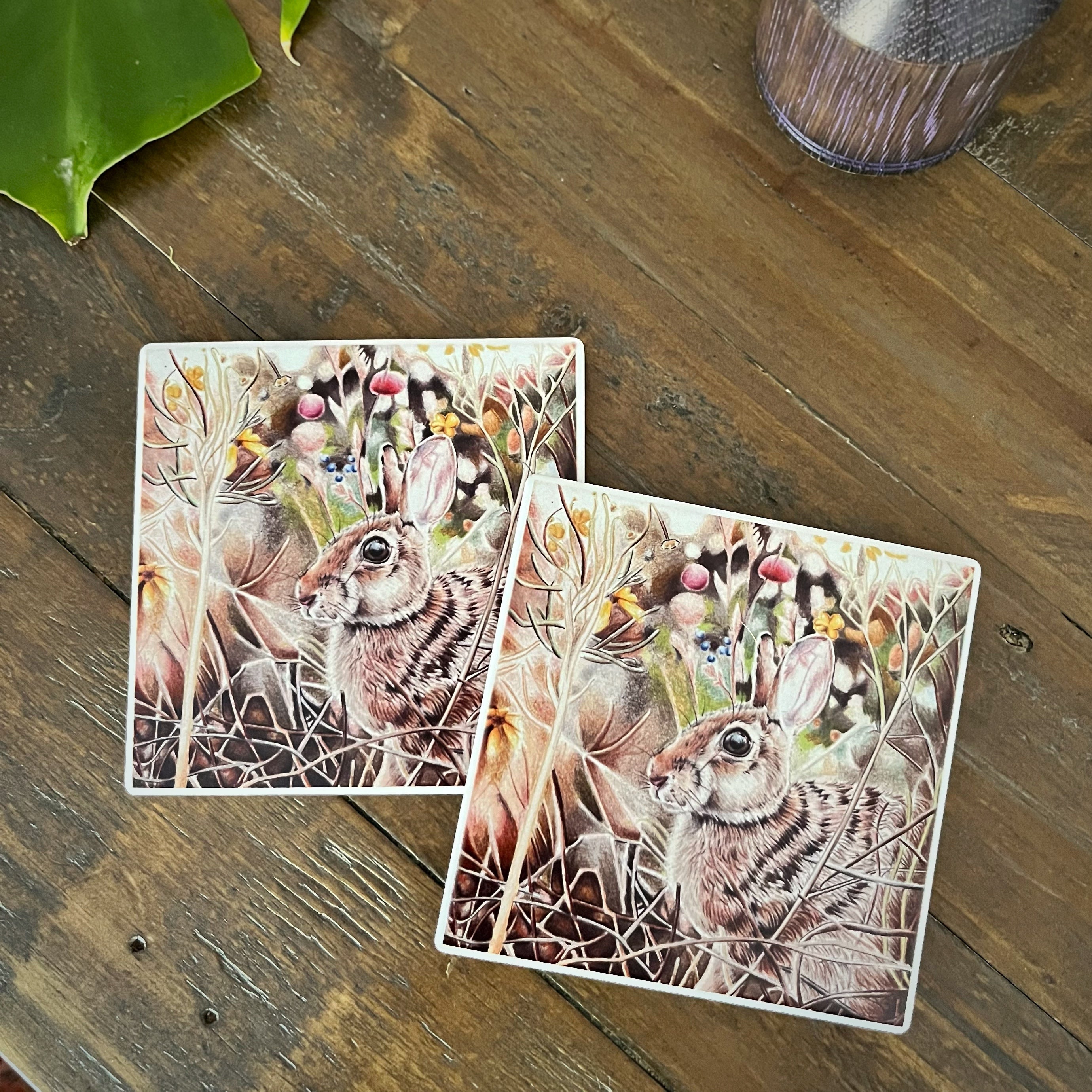 Two square coasters with a detailed illustration of a rabbit in a field of grass and wildflowers, displayed on a rustic wooden tabletop.