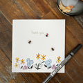 A square white card with a watercolor-style floral and bee design, placed on a wooden tabletop with a black pen resting across the lower right corner.