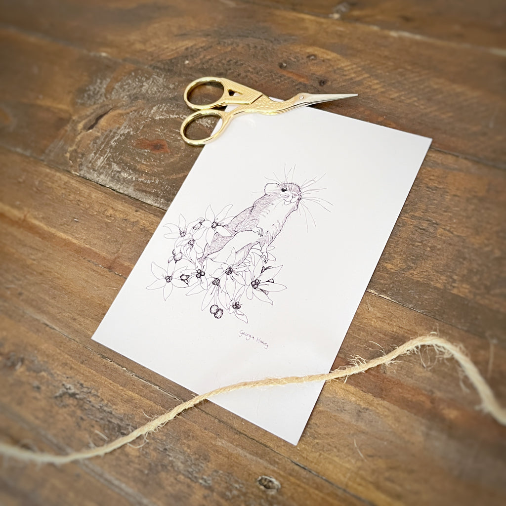 A white art print featuring a delicate pen sketch of a small field mouse among flowers, styled on a rustic wooden table with gold-handled scissors and twine