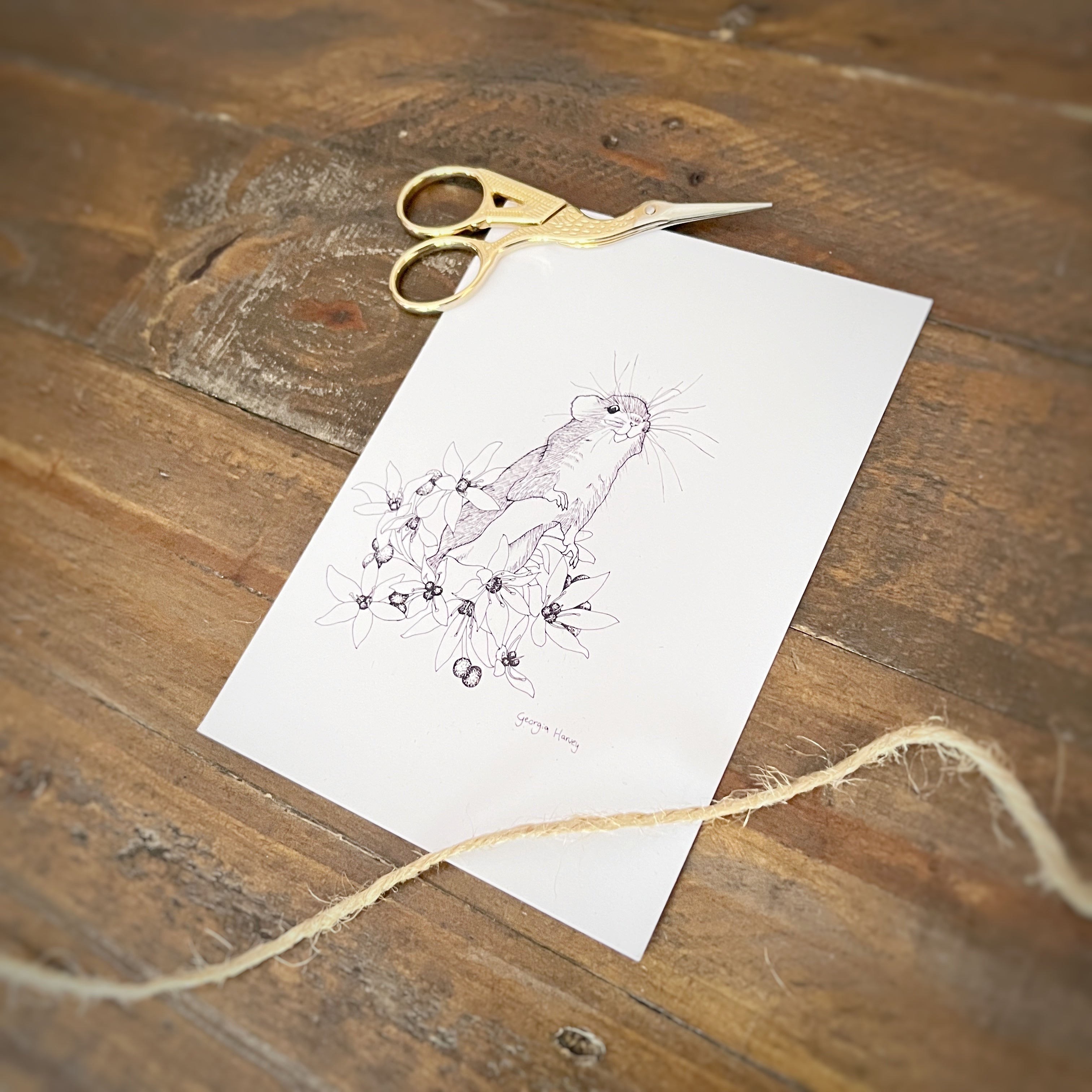 A white art print featuring a delicate pen sketch of a small field mouse among flowers, styled on a rustic wooden table with gold-handled scissors and twine