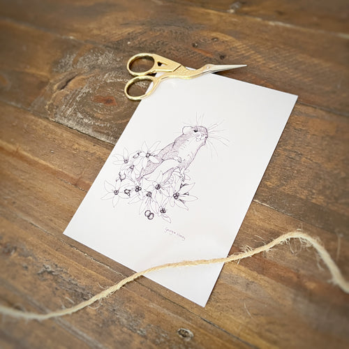 A white art print featuring a delicate pen sketch of a small field mouse among flowers, styled on a rustic wooden table with gold-handled scissors and twine