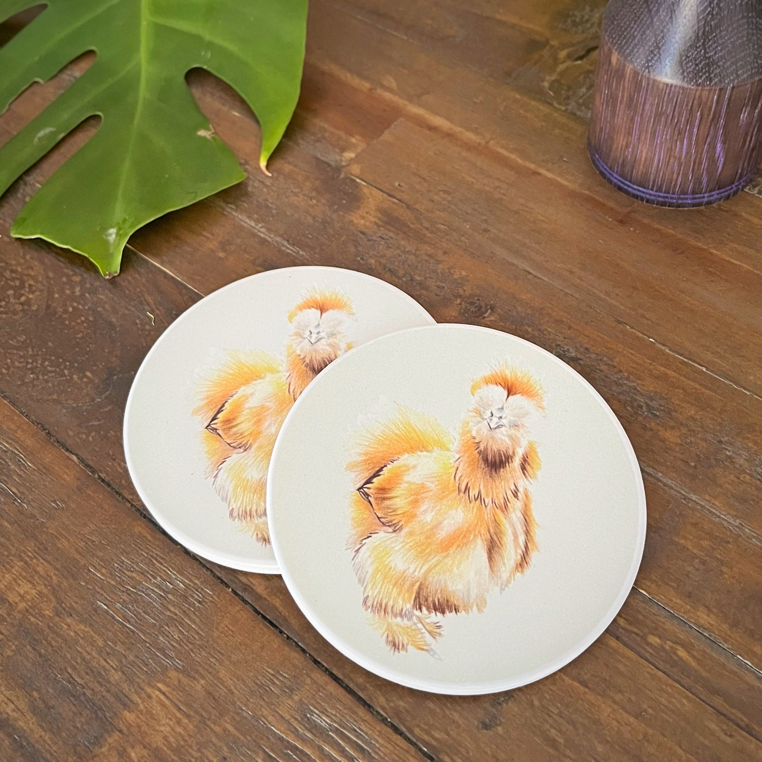 Two round coasters featuring a watercolor-style illustration of a fluffy golden silkie chicken, displayed on a dark wooden tabletop next to a green leaf and a glass container.