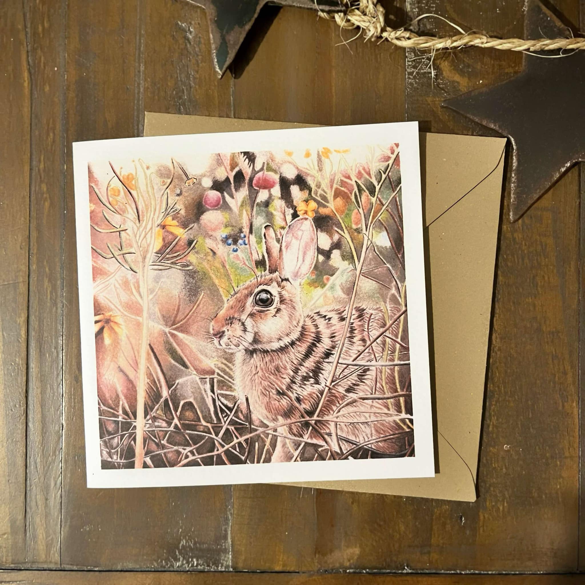 A square greeting card featuring a detailed illustration of a brown rabbit among autumn foliage, displayed on a rustic dark wood surface with a kraft envelope behind it.