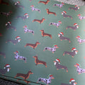 Green wrapping paper featuring a repeating pattern of dachshund sausage dogs wearing red Santa hats, photographed flat with a slight angle 