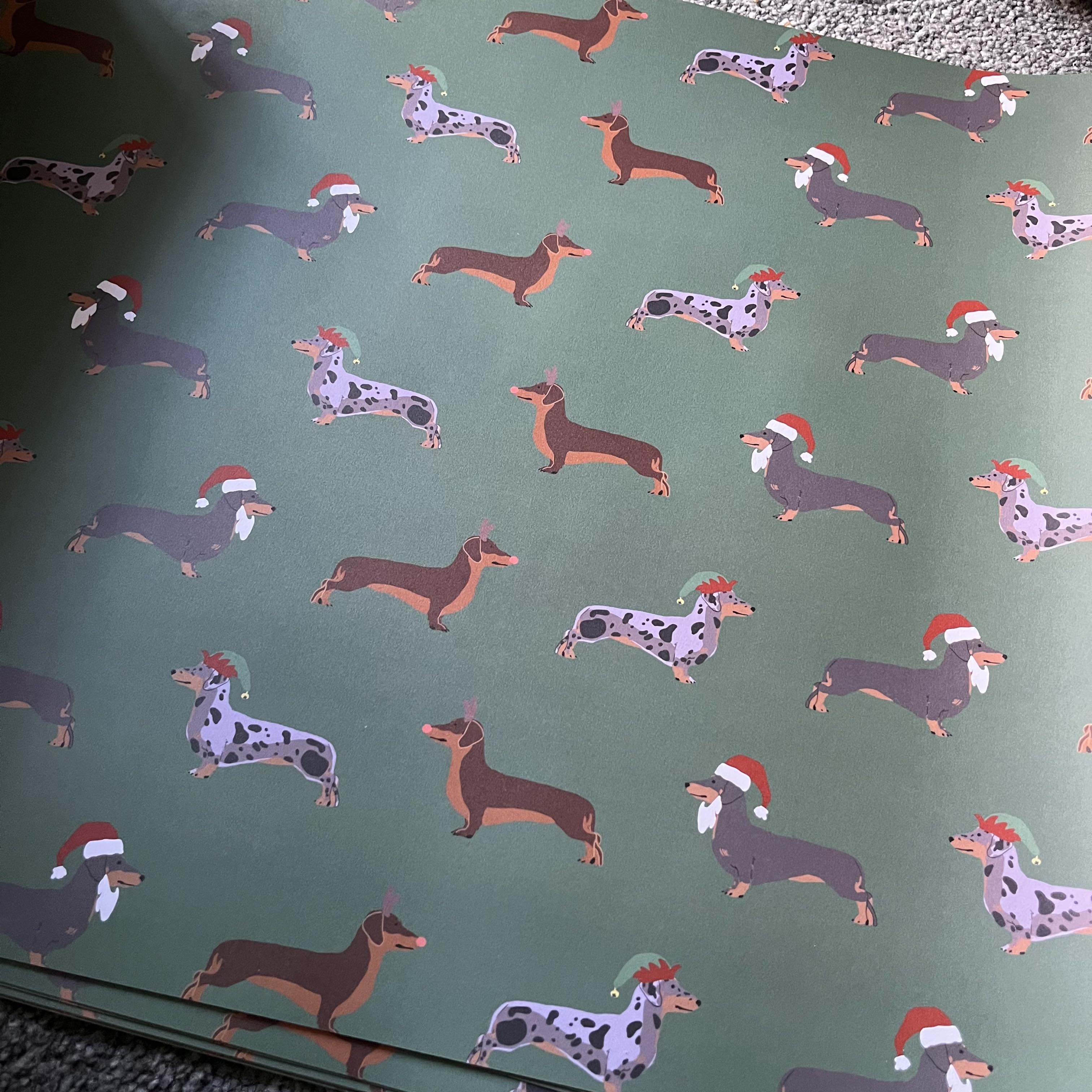 Green wrapping paper featuring a repeating pattern of dachshund sausage dogs wearing red Santa hats, photographed flat with a slight angle 
